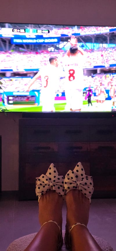The image captures a person's feet wearing polka dot slippers with bows, resting on a textured surface, in front of a television. The television screen displays a soccer match, possibly from the FIFA World Cup 2018, with players in white jerseys and a scoreline indicating 0-0. The background of the television screen shows a stadium filled with spectators. The overall scene suggests a relaxed atmosphere, perhaps at home, watching a sporting event. There is no discernible information about the city of Lieli, Switzerland, in this image.