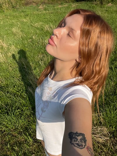A young woman with ginger hair and closed eyes tilts her head upwards, basking in sunlight. She wears a white t-shirt with a subtle floral print. A cartoonish tattoo of a face is visible on her right forearm. The background consists of a grassy field under a clear sky, casting a shadow of the woman onto the ground. The scene appears to be outdoors, possibly in a park or rural area, given the natural landscape.