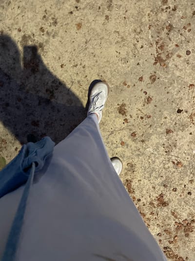 A first-person perspective shot shows a person standing on a concrete surface. The person is wearing light blue pants and white sneakers. A blue bag is visible on the left side of the frame. A shadow of the person, as well as a shadow of what appears to be a phone, is cast on the ground. The concrete surface is speckled with small brown debris. The scene is outdoors and appears to be a public area in La Línea De La Concepción, Spain.