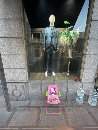 The image shows a storefront window display. Inside the window, a mannequin is dressed in a suit with a black tie and white shirt. White flowers are arranged behind the mannequin. To the right of the window, a pink toy stroller with a Hello Kitty design is visible, along with two plastic water bottles. The storefront is made of stone blocks, and the ground is paved. A metal railing is in the foreground. The scene appears to be set during the day, with natural light reflecting on the glass. There are no visible signs of weather conditions. The setting suggests a commercial area, likely a clothing store or boutique. The image is oriented sideways.
