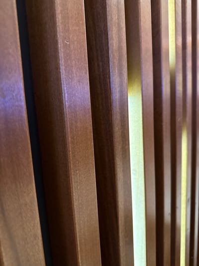 The image shows a close-up, angled view of parallel, horizontal wooden slats with light shining through the gaps between them. The wood is a medium brown color. The perspective suggests the viewer is looking upwards at the slats. The scene appears to be an architectural element, potentially a wall covering or screen. There is no evidence in the image to suggest the location in Hollywood Seminole Reservation, United States of America. The image contains no people, subjects, or actions.