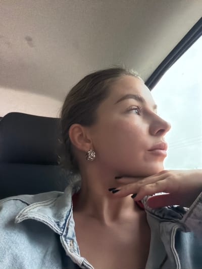 A young woman is shown from the chest up, in profile, looking out of a car window. She has fair skin, dark eyebrows, and dark eyelashes. Her lips are slightly parted, and she has her right hand resting on her chin. She is wearing a light blue denim jacket over a red top. Her fingernails are painted black. She has a silver earring in her right ear. The interior of the car is visible behind her, with a dark seat and the grey headliner of the car. The scene appears to be a bright, overcast day. The location is not identifiable as Gornji Vakuf-Uskoplje.