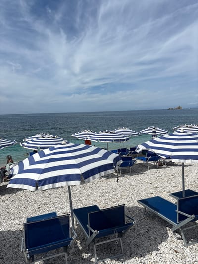 This image captures a picturesque beach scene in Portoferraio, Italy, under a partly cloudy sky. The setting features a pebble beach adorned with numerous blue and white striped beach umbrellas, mostly open, stretching along the shoreline. Accompanying the umbrellas are blue beach chairs and loungers, some in use, others folded.

The calm, blue-green sea extends to the horizon, where it meets a sky streaked with soft, wispy white clouds. A distinct rocky outcrop or small island is visible in the distance on the left side of the sea. A few blurry figures of people are discernible among the umbrellas, suggesting the beach is in use, with one individual's head and shoulder visible behind an umbrella in the foreground.

The time of day appears to be late morning or early afternoon, evidenced by the bright sunlight and the shadows cast by the umbrellas and chairs on the white pebbled sand. On one of the blue beach chairs, partially visible white text is present, though it is too indistinct to be clearly deciphered. The scene conveys a relaxed, vacation-like atmosphere.