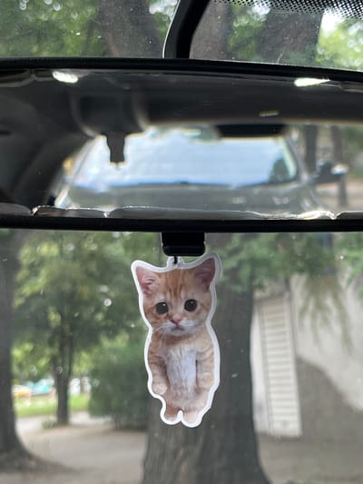 An orange tabby kitten air freshener hangs from a car's rearview mirror. The kitten is depicted as a cartoonish sticker with large eyes and a pink nose. The background appears to be a residential street with trees and buildings, suggesting an outdoor setting in a city or town. The interior of the car is visible in the reflection of the mirror, showing a glimpse of the dashboard and windshield. The image was captured in Orizari, Bulgaria.