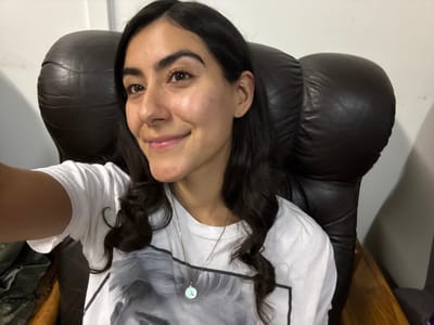 A young woman with long, dark, wavy hair is sitting in a brown leather chair, facing the camera. She is wearing a white t-shirt with a black and white image printed on it and a gold necklace with a light blue pendant with the letter "A" on it. She is smiling slightly. The background appears to be an indoor setting, with a white wall and part of a dark object to her left. There is no indication that this image was taken in Imatra, Finland.