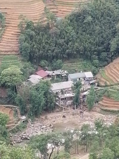 Cat Cat Village, Vietnam. A high-angle view shows a terraced landscape with buildings nestled amongst trees and a rocky riverbed. Terraced fields are visible on both sides of a valley, with varying levels of vegetation and cultivated areas. Structures with roofs of gray and red are interspersed among the foliage. Construction is also apparent. Below the structures is a wide, rocky area, possibly a dry riverbed. Trees and other greenery line the foreground, partially obscuring the view. The overall impression is a rural landscape with terraced farming and scattered human settlements.