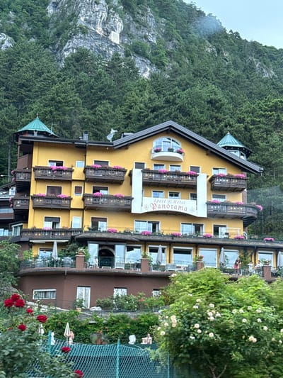 The image shows a yellow hotel named "Sport Hotel Panorama" nestled on a mountainside in Fai della Paganella, Italy. The hotel has multiple levels with balconies adorned with pink flowers. A dense green forest covers the steep mountain behind the hotel. In the foreground, lush greenery and red roses are visible, along with a teal chain-link fence. Several people are present on the hotel's balconies and terraces. The overall scene depicts a scenic mountain resort.
