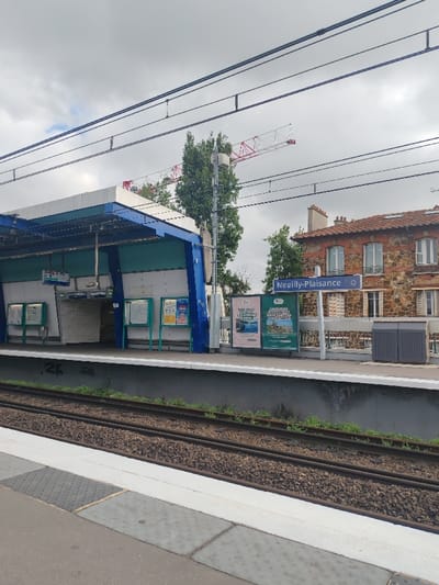 The image depicts a railway station platform under an overcast sky in Neuilly-Plaisance, France. In the foreground, two parallel train tracks are visible on a bed of gravel ballast, running alongside a concrete platform. The platform edge features a white safety line and grey tactile paving.
On the platform, a modern station shelter with a blue structural frame and a white roof is present. Its interior walls are a light blue-green color, and it contains openings suggesting access to a lower level or underpass. Several information or advertising panels with green frames are mounted on the platform wall beneath the shelter. To the right, a blue sign with white lettering clearly displays "Neuilly-Plaisance" and a circular symbol. Adjacent to this sign are two grey, lidded waste bins.
In the background, a large red and white construction crane boom is visible above tall green trees. To the right, a multi-story building with a red tiled roof and a stone-textured or brick facade, featuring several windows with light-colored frames, stands behind the platform. The sky is uniformly grey and cloudy. No individuals are present in the scene.