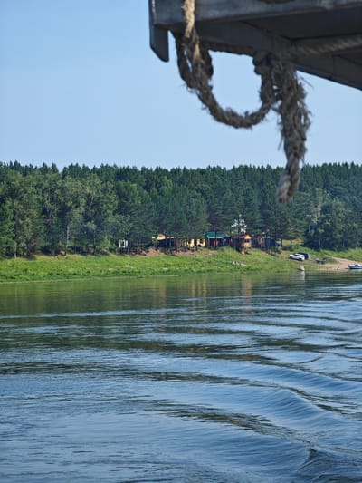 The image captures a serene river scene in Podporozh'e, Russia, on a clear, sunny day. In the foreground, the gentle ripples of the water create a dynamic surface. The middle ground features a lush, green riverbank densely populated with a variety of trees, predominantly evergreens, with a few deciduous trees interspersed. Along the riverbank, a small settlement of colorful buildings, possibly cabins or cottages, is visible amongst the trees. Several vehicles, including a white SUV and a trailer, are parked near the shore, along with a small boat. The background consists of a clear, light blue sky. A thick, weathered rope hangs from the top right of the frame, partially obscuring the sky and the upper edge of the riverbank. The overall impression is one of a peaceful natural setting with signs of human habitation.