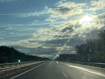 The image shows a view from a highway. The highway is paved and has white lines marking the lanes. On the left side of the highway, there is a metal guardrail, and beyond that, a densely vegetated slope. On the right side, there is also a guardrail and a wooded area. The sky is filled with clouds, with the sun breaking through in the upper right portion of the image, creating lens flares. There are contrails from airplanes visible in the sky. In the distance, there are several vehicles on the highway. The scene appears to be a rural or suburban setting, possibly in or near Kolarovo, Bulgaria, given the context provided.