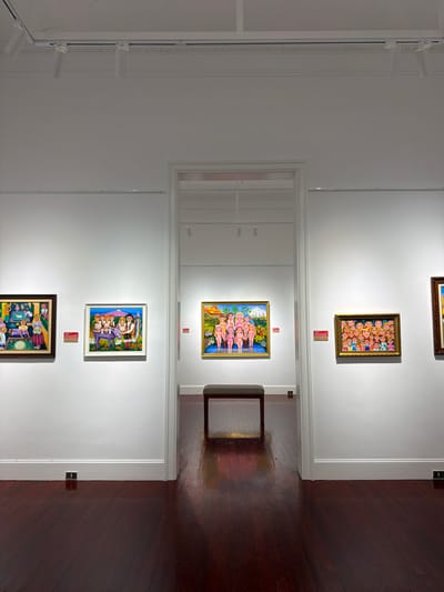 This is a wide shot of an art gallery with polished dark wood floors and white walls. Several paintings are displayed on the walls. On the left, a framed painting depicts a group of people, possibly a family, with a lamb and religious imagery. Next to it, another framed painting shows a family, including a child on a donkey, surrounded by sunflowers with a mountain in the background. In the center, through an open doorway, a bench is placed in front of a painting of a group of nude figures standing in water. To the right of the doorway, another framed painting displays a crowd of people with exaggerated facial features. Further to the right, a sliver of another framed painting is visible. Track lighting is installed on the ceiling.