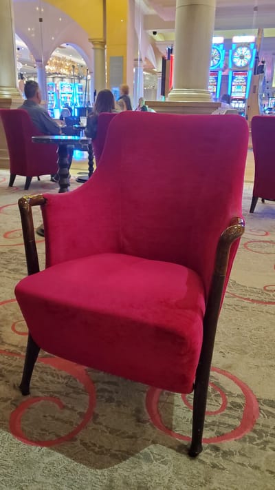 The image captures a close-up view of a plush red armchair in the foreground, positioned on a patterned carpet. The armchair has dark wooden armrests with a curved, polished finish. In the background, blurred figures of people are visible, and what appear to be slot machines with illuminated screens suggest the setting is a casino, likely within Atlantic City, United States of America. Architectural elements like columns and arches, as well as the general ambiance, further support this identification of a casino environment. The focus is primarily on the armchair, with the background elements providing context.