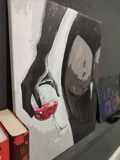 The image shows a close-up of a painting, likely acrylic on canvas, displayed on a gray shelf. The painting depicts an abstract, minimalist representation of a human form holding a glass of red liquid. The colors used are primarily dark browns, grays, white, and vibrant red. To the left of the painting are two books, one with a red cover and another with a dark cover adorned with red floral patterns. Behind the painting and books, a dark gray textured wall is visible. To the right of the painting, a smaller piece of art, possibly a print or another painting, featuring colorful handprints, is placed. A black electronic device is also visible to the right of the smaller artwork. The scene appears to be an indoor setting, possibly a home or gallery. There is no indication of the city of Reftinsky, Russia within the image itself.