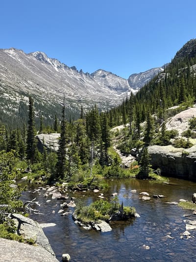 This image depicts a natural mountain landscape with a clear sky. In the foreground, a shallow body of water with a rocky bottom is visible, with several larger rocks and small islands of vegetation scattered within it. The water is edged by a rocky shore with some sparse green vegetation.

Dominating the mid-ground and background are steep, rocky mountainsides covered in dense evergreen trees. The mountains themselves are rugged and appear to be made of granite, with patches of snow still visible in shaded areas and crevices, indicating a high altitude and possibly a recent thaw. The peaks of the mountains are sharp and jagged, and the overall impression is one of a wild and remote natural environment.

The scene is illuminated by bright sunlight, casting distinct shadows. The overall color palette is dominated by the blues of the sky, the grays and whites of the mountains, and the greens of the coniferous forest.

While this image clearly depicts a mountain environment, there is no specific visual information that allows for definitive placement within the city of Estes Park, United States. However, the type of landscape is consistent with national parks and wilderness areas found in and around mountainous regions like Estes Park, which is known for its proximity to Rocky Mountain National Park. Therefore, it is plausible that this scene is located in such a mountainous region. There are no people or human structures visible in the image.