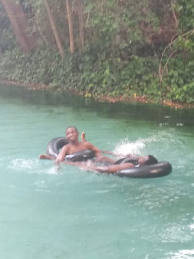 Two individuals are floating on black inner tubes in a body of water that is light green. The person on the left is smiling and giving a thumbs-up. The person on the right appears to be lying back. The background consists of dense green vegetation and some tree trunks. The location is Lim Kundak, Nigeria.