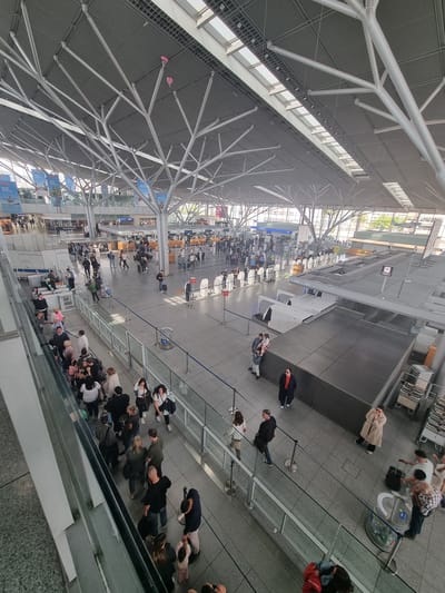 The image depicts the interior of Stuttgart Airport (STR) in Echterdingen, Germany, from an elevated perspective. The airport features a high ceiling with a complex network of steel beams and skylights. The floor is gray and tiled, with multiple queues of people waiting in line, presumably for security or check-in. Some individuals are seated while others are standing. Baggage carts and conveyor belts are visible in the background. Glass barriers or partitions partially separate the queueing areas. Signage is visible, indicating various terminals and gates. The overall scene suggests a busy airport environment with numerous travelers.