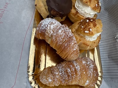 The image displays a collection of Italian pastries on a gold-colored tray. The pastries consist of at least two Sfogliatelle, dusted with powdered sugar, and several cream puffs, some filled with white cream and adorned with sugar pearls, and one with a chocolate glaze. The pastries are presented on a corrugated gold-colored tray. The tray rests on a surface covered with printed white paper with stripes and a red line and text are visible on the right. Based on the paper, this image was taken in Tivoli, Italy.