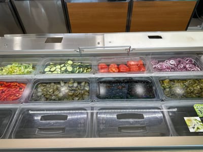 The image displays a refrigerated salad bar at a Subway restaurant, likely located in Mikhailovka, Russia, based on the Cyrillic labeling on some of the containers. The salad bar is filled with various fresh ingredients, including chopped lettuce, sliced cucumbers, sliced tomatoes, sliced red onions, sliced red bell peppers, pickle slices, black olives, and sliced green jalapeños. The ingredients are arranged in clear plastic containers, which are set within a larger stainless steel refrigerated unit. The lighting from above illuminates the fresh ingredients, highlighting their vibrant colors. Below the ingredient containers, there are empty clear plastic trays, suggesting that the salad bar is ready to be served. In the background, a stainless steel counter and wooden cabinetry are visible, typical of a fast-food establishment. The scene is clean and organized, indicating a well-maintained food service environment.