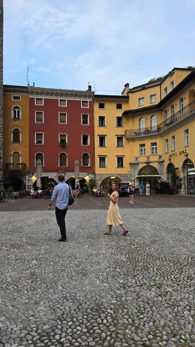 The image captures a lively scene in a European town square. The main subjects are a man and a woman, both depicted in mid-motion. The woman, wearing a light yellow dress and sandals, appears to be twirling or dancing, her movement creating a dynamic line across the cobblestone ground. Her expression is unreadable but her pose suggests joy or abandon. The man, dressed in a blue shirt and black pants, is shown in a low crouch, as if about to push off or in the process of kneeling. He carries a dark bag over his shoulder.

The setting is an outdoor plaza paved with smooth, grey cobblestones. To the left, a row of colorful buildings rises, painted in shades of yellow and red. These buildings feature numerous windows, balconies with ornate railings, and awnings. One building prominently displays signage that reads "HOTEL SOLE" in a yellow illuminated sign, and beneath it, "HOTEL SOLE". Another sign, partially visible, appears to be for a cafe. The architecture suggests a historic European town. In the background, more people are visible, some seated at tables under umbrellas, indicating a cafe or restaurant. The sky is a bright, clear blue with faint white clouds, suggesting a pleasant daytime. The overall atmosphere is one of activity and casual enjoyment, possibly during a warm afternoon in Riva del Garda, Italy, as hinted by the location context.