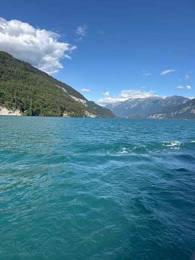 The image captures a serene view of Lake Thun from a boat, looking towards the mountains beyond Sigriswil, Switzerland. The foreground is dominated by the vibrant turquoise water of the lake, with gentle ripples and small waves suggesting movement. In the midground, a densely forested green mountain slopes steeply down to the water's edge, with a small village nestled along the shoreline, identifiable by scattered buildings and structures. The background features a majestic range of rugged, grey mountains under a clear blue sky dotted with fluffy white clouds. The overall impression is one of natural beauty and tranquility.