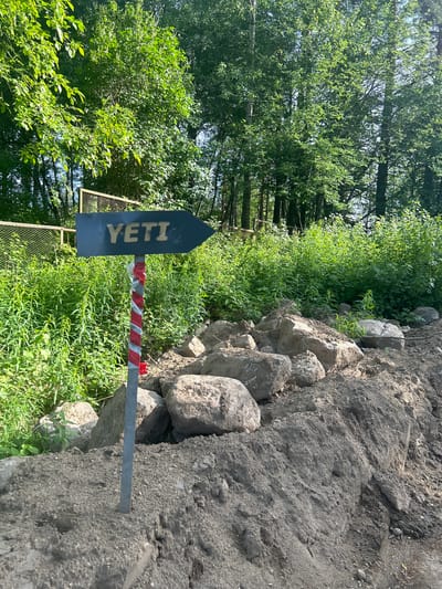 A blue directional sign with the word "YETI" in white letters points to the right. The sign is mounted on a thin gray metal pole. Red and white striped tape is wrapped around the pole below the sign. The pole is embedded in a pile of dirt and rocks. Lush green foliage and trees are visible in the background. A chain-link fence can be seen behind the foliage. The image appears to be taken outdoors during the daytime.