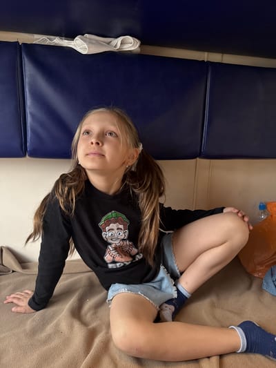 A young, fair-skinned girl with blonde hair is sitting on a neutral-toned surface resembling a blanket or couch. She is wearing a black long-sleeved shirt with a cartoon graphic on the front and blue denim shorts. Her legs are bent, and she is wearing socks. To the left of the frame, there is a blue padded surface, possibly a seat or wall. In the background, there are miscellaneous items including a plastic bag and another piece of clothing. The location is potentially Volkhov, Russia.