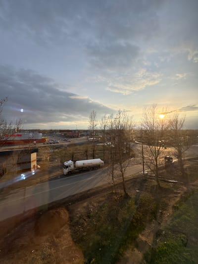 The image shows a view over Kladovitsy, Russia, featuring a cloudy sky with a sunburst effect peeking through bare trees. A road runs through the middle ground, where a white truck with a cylindrical tank trailer is parked. Further along the road, there are parked cars and what seems to be commercial buildings, possibly a store, with a partially constructed structure at the lower edge. The immediate foreground is blurry, perhaps the inside of a window. The right side of the image shows a patch of grassy or barren land.