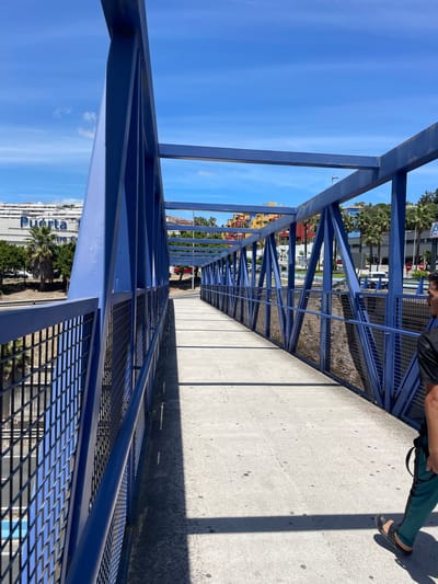 The image shows a person's legs and feet from the knees down, wearing dark green pants and black sandals. The person is standing on a concrete walkway of a blue metal pedestrian bridge. The bridge has a latticed metal railing. The scene is outdoors on a sunny day, casting shadows on the walkway. In the background, there are palm trees, colorful buildings (yellow and red), a street with cars, and a large white building with text that appears to read "PUERTA" which is Spanish for "door" or "gate". The sky is clear and blue. The orientation of the image is sideways.