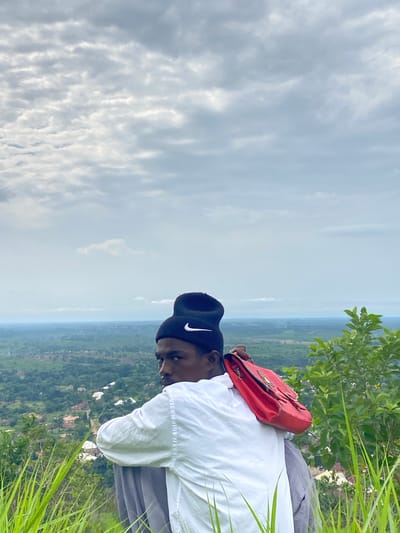A young man wearing a black beanie with a white Nike logo and a white long-sleeved shirt is sitting on a grassy slope overlooking a landscape. He is holding a red handbag over his shoulder. The background shows a hazy view of a town with scattered buildings and lush green vegetation, characteristic of the area around Ufu Esioni, Nigeria. The sky is overcast with layered clouds. Tall, green grass is in the foreground, partially obscuring the lower part of the man's body.