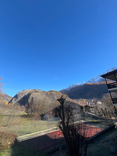 This image displays an autumnal, panoramic view of a mountainous landscape in La Thuile, Italy, under a clear, deep blue sky. No individuals, discernible actions, or interactions are visible.

The scene encompasses a foreground featuring a red-surfaced sports court, likely a tennis court, marked with white lines and surrounded by green fencing. Adjacent to the court, to the left, is a grassy area dotted with bare deciduous trees and some low-lying shrubbery. A light-colored van is parked among trees further back on the left side.

In the midground, a collection of traditional alpine buildings with terracotta roofs is clustered, indicating a village section. Directly to the right, partially framing the image, is the dark-colored roof and wooden balcony of a building, likely the vantage point of the photograph.

The background is dominated by mountains. To the left, a large, rocky mountain face is visible, sparsely covered with bare trees and patches of yellow-orange autumn foliage. To the right, a higher mountain range features snow-capped peaks, with the lower slopes covered in dense coniferous forests displaying vibrant golden and orange autumn colors. The lighting is bright and consistent with a sunny day.