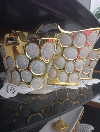 This image captures an indoor display in Nasiriyah, Iraq. Three decorative containers, gold-toned with silver-glittered circular inlays, are arranged on a white lace doily atop a dark, highly reflective surface, likely glass. The containers decrease in size from left to right; the center container is fully visible, while the one on the far left and the one on the far right are partially cropped. Each container features integrated handles. Below this display, a lower shelf holds a white rectangular dish featuring a small, golden, domed object on a stand, partially resting on another white lace doily. The background is dark and indistinct, with a suggestion of green foliage visible on the far right. No individuals are present, and the objects are stationary.