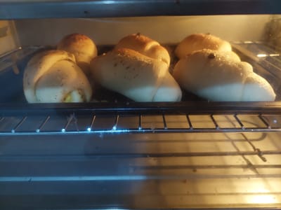 Six crescent-shaped baked goods, possibly rolls or pastries, are arranged on a black baking tray within an oven. The baked goods have a light brown exterior with some darker specks, possibly herbs or spices, sprinkled on top. The oven's interior is illuminated by a warm yellow light. The baking tray is placed on a metal rack, and the glass door of the oven is visible in the foreground. The image appears to have been taken from a slightly elevated angle, looking down into the oven. The scene is devoid of any human presence or activity. There are no discernible landmarks or contextual elements to place this image within the city of Jelutung, Indonesia.
