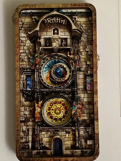The image displays a miniature, three-dimensional diorama set within a rectangular frame, reminiscent of a shadow box or a decorative tin. The diorama depicts a detailed architectural scene with a strong emphasis on mechanical elements, suggesting a clock tower or a similar intricate structure.

The central focus is on two circular, clock-like mechanisms. The left mechanism features intricate blue and gold spiraling patterns, while the right one is adorned with a yellow and red ornamental design. These are framed by a backdrop of textured, brick-like patterns in shades of brown and beige, giving the impression of an aged stone building. Small, colorful figures, possibly representing people or characters, are strategically placed around the mechanisms and along the architectural elements, suggesting they are interacting with or observing the structure. Some of these figures appear to be in motion.

Above the clock mechanisms, there are small framed images or windows displaying what appear to be portraits or scenes. To the left of the clocks, a curved architectural element prominently displays the word "PRAHA" in an ornate font, indicating Prague as the likely inspiration for the diorama. The overall aesthetic is complex and detailed, with a slightly weathered or antique appearance due to the color palette and texture.

The scene is indoors or presented as a static display, with no clear indication of time of day or weather. The environment is highly stylized and imaginative. The emotion conveyed is one of wonder and appreciation for intricate craftsmanship.