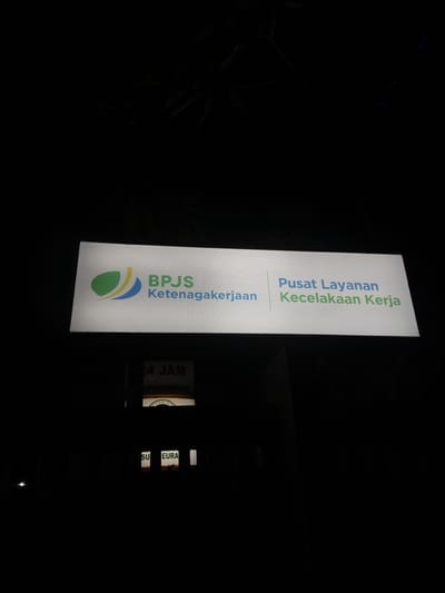The image shows a lit sign at night in Lampeuneurut Blang, Indonesia. The sign features the BPJS Ketenagakerjaan logo, which consists of green and blue curved shapes. The text on the sign reads "BPJS Ketenagakerjaan" and "Pusat Layanan Kecelakaan Kerja" in blue and green lettering. Below this main sign, another smaller illuminated sign is visible with text that includes "24 JAM." Further down, another sign has the partial text "SU EURA". The surrounding environment is dark, suggesting it is nighttime, with some branches of a tree visible against the sky at the top of the image. The image is captured from a slightly low angle, looking upwards at the signs.