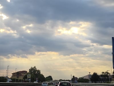 The image depicts a highway scene with a dramatic sky filled with heavy clouds and shafts of sunlight breaking through. Several cars are visible on the highway, suggesting traffic. On the left side of the road, there are buildings, streetlights, and trees. A speed limit sign indicating 50 is visible on the right side. The overall atmosphere is overcast with pockets of bright light. The location is identified as Carinaro, Italy.