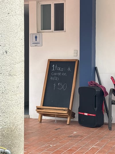 The image presents an outdoor scene adjacent to a light-colored building in San Francisco Totimehuacán, Mexico. A window is visible on the building's wall along with a men's restroom sign ("HOMBRES"). Adjacent to the restroom is a chalkboard sign, propped on a wooden easel, advertising "1 taco de carne en tiras $50" (1 taco of sliced meat $50). A black KSR speaker, partially covered with a dark cloth, sits on the ground and appears to be supporting the easel. The ground is divided between textured concrete paving in the foreground and brick paving in the background.
