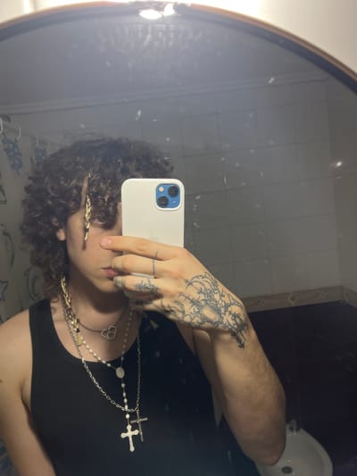 A person is taking a selfie in a bathroom mirror. The person is wearing a black shirt and multiple necklaces, including one with a cross. They have curly hair and braids on the side of their face. Their left hand is holding a white smartphone up to their face, obscuring their nose and part of their eyes. The left arm has a tattoo. The bathroom has white tiled walls, a dark countertop, and a white sink visible in the upper right corner. The image does not contain information regarding the city of Bahía Blanca.