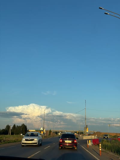This image depicts a view from a vehicle traveling on a highway in Derzhavino, Russia. The sky is a clear, bright blue with a few wispy clouds scattered across the upper portion, and a large, towering cumulus cloud formation is visible on the horizon. The road is paved and has lane markings. In the foreground, two cars are on the highway. The car closest to the viewer is a dark red sedan with license plate E125OK716. Ahead of it is a white sedan with a license plate that appears to read B667HH49RUS. The road is lined with grassy verges and trees. In the distance, there are streetlights, overpasses, and construction barriers, indicating ongoing roadwork or infrastructure development. The overall scene suggests a summer day with good visibility.