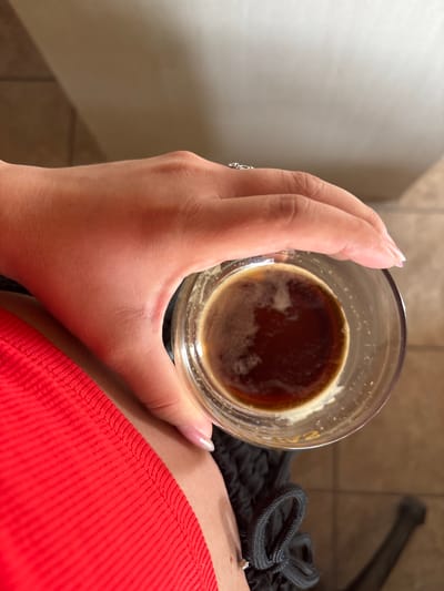 A close-up, high-angle shot captures a person holding a clear glass containing a dark brown beverage, likely coffee, with a layer of light foam or crema on top. The subject's hand, with a light skin tone and manicured nails featuring a light polish, is prominent in the center-left, gently gripping the glass. A silver-colored ring with a small, clear gemstone is visible on the ring finger.

The person is wearing a vibrant red, ribbed fabric garment, partially visible on the right side of the frame, and a black garment with a drawstring, which appears to be shorts or sweatpants, just below the glass.

The background, partially visible on the left and upper right, consists of neutral-toned tiled surfaces, possibly a floor and a lower wall section, indicating an indoor setting. A blurry dark object is present in the upper right background. The image focuses tightly on the hand and drink, with the overall scene being well-lit. The activity depicted is simply holding a drink. No specific time of day or emotions are discernible, and no text is visible in the image. The scene is located in Orizari, Bulgaria.