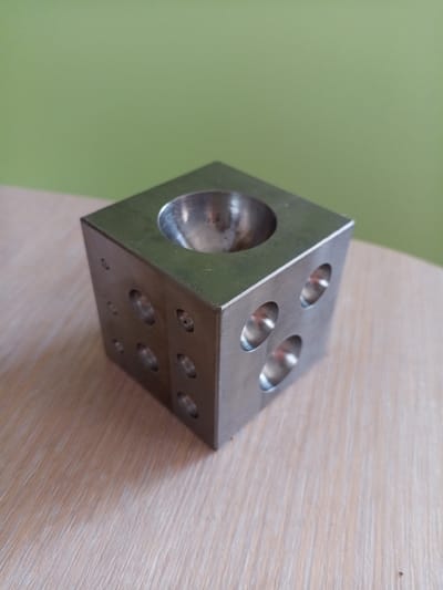 A metallic cube with multiple circular indentations on its sides and a hemispherical indentation on its top surface rests on a light-colored wooden table. The background is a solid, light green wall. The lighting is even, with no harsh shadows. The image is a close-up of the object, highlighting its metallic texture and the precision of its machined indentations. The location is Vilyuysk, Russia.