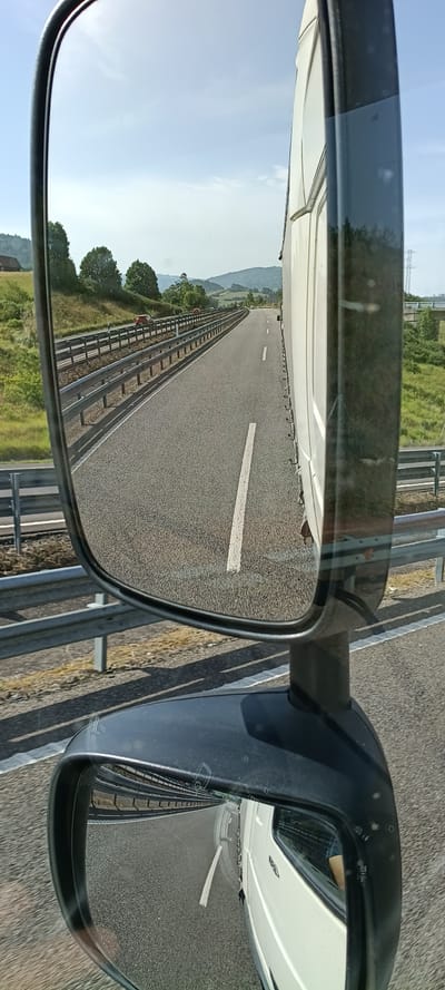This media file depicts a view from inside a vehicle, looking out through its side mirrors. The primary scene is a highway in Villaviciosa, Spain. The road is paved with asphalt and features a white dashed line marking the lanes. A metal guardrail runs parallel to the road. In the distance, rolling green hills and trees suggest a rural or semi-rural setting. The sky is a clear, light blue with a few wispy clouds, indicating fair weather during daylight hours. The time of day appears to be morning or afternoon, given the brightness of the sky.
The subjects are the highway itself, the vehicle's side mirrors, and the surrounding landscape. The activity taking place is travel along the highway. Notable details include the reflections in the mirrors, which show more of the road and the vehicle's white bodywork. The perspective is from a slightly elevated position, typical of a truck or van. No people are clearly visible, though there might be indistinct figures in the distance on the highway. There is no visible text in the image. The overall impression is one of a journey through a scenic landscape.