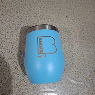 A light blue, stemless wine tumbler with a metallic rim is placed on a speckled beige countertop. The tumbler features a metallic logo on the side, with the word "BYOND" inside a rectangular outline and a large stylized letter "B" to the right. Below this, the text "by BSi" is also visible. The background is out of focus, showing the texture of the countertop. There are no people present in the image. The image appears to have been taken indoors, possibly in a kitchen or dining area in Reudeuep, Indonesia.