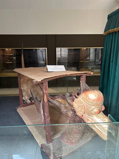 The image shows a display inside a museum. The central object is a wooden structure that appears to be a throne or a palanquin, with a woven mat for seating and a carved wooden head of a mythical creature, likely a Garuda, at the front. The creature's head is intricately decorated with red and gold paint. A sign on the seating mat reads "TANDU GARUDA MINA." In the background, display cases with various artifacts are visible, some containing what appear to be weapons or tools. A dark green curtain with gold trim hangs on the right side of the frame. The entire display is protected by a glass barrier. The context provided indicates this is located in Cirebon, Indonesia, a city known for its historical sites and cultural heritage, including its palaces and museums which often house traditional Javanese art and artifacts.