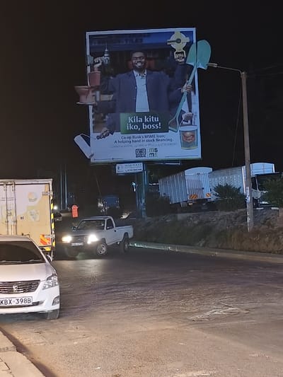The image depicts a night street scene with various vehicles, including a white sedan with license plate KBX-398B and a white pickup truck. Above the road is a large billboard advertising Co-op Bank's MSME loan, featuring a man in a suit surrounded by tools and construction materials. The billboard reads "Kila kitu iko, boss!" and includes contact information for the bank. Further up the road, several large trucks are parked or moving.