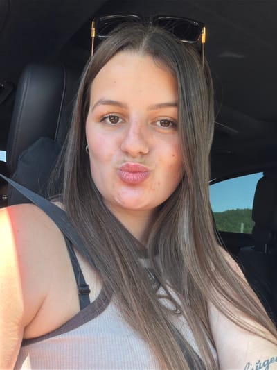 A young woman is taking a selfie in a car. She has long, dark brown hair with some lighter streaks. She is wearing sunglasses on her head and a grey top with a black bra visible underneath. Her lips are pursed, and she has a few freckles on her cheeks. The car interior is visible, including a black leather seat and a seatbelt. Sunlight is visible on her shoulder and arm, suggesting it is daytime. The background outside the car window appears to be trees. The image is oriented vertically. There is no visible text.