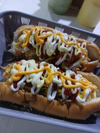 The image shows two hot dogs in a gray plastic tray. The hot dogs are topped with french-fried potatoes, mayonnaise, ketchup, and a yellow cheese sauce. The food is set on a white surface. A container of yellow sauce is visible on the left edge of the image. The location is El Espinal, Venezuela.