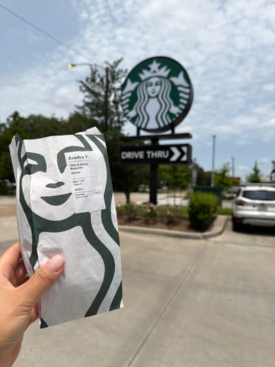 A person holds a Starbucks bag with a "Ham & Swiss Baguette" receipt visible. The receipt indicates the order was placed on June 25, 2026, at 12:44 PM via mobile ordering and was item 1 of 1. The bag is white with Starbucks' signature green wavy pattern. In the background, a Starbucks sign with the iconic siren logo is visible, along with a "Drive Thru" sign. The scene appears to be outdoors, likely a bright day with some clouds, suggested by the lighting and sky. A car is partially visible in the distance, and there are trees and greenery around. The overall impression is of a casual transaction at a fast-food establishment.