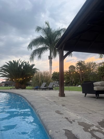 The image shows an outdoor scene at dusk or dawn in Santa Ana Yancuitlalpan (Chilhuacán), Mexico. The view is from under a dark, wooden structure, possibly a gazebo or pergola. A tall palm tree extends upwards toward a sky with a mix of grey clouds and a strip of orange-tinted sky, suggesting either sunrise or sunset. A second, shorter palm tree is decorated with string lights. To the right, a patio area is visible, with outdoor furniture including chairs and cushions. In the foreground, the edge of a blue-tiled swimming pool is visible. In the background, additional trees are strung with lights. The patio area appears wet, suggesting recent rain. There are no people visible in the scene.