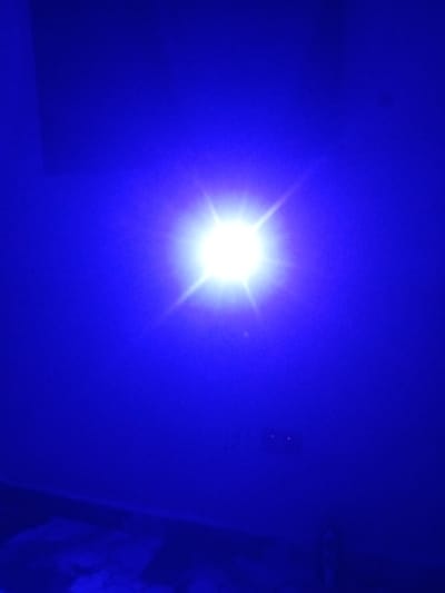 The image shows a bright white light source with starburst effects against a deep blue background. The light source is positioned slightly off-center, to the left of the vertical midline of the image. The blue background appears to be a wall, with some subtle variations in shade suggesting uneven lighting or texture. In the lower right portion of the image, two small, red illuminated elements are visible, possibly indicator lights on an electrical outlet or device, suggesting an indoor environment. The bottom of the image shows a dimly lit, textured surface that could be a floor with some patterned elements. The overall impression is that of a photograph taken in a dark room illuminated by a strong, focused light source. The location in Tundun Wada West, Nigeria, does not provide any further visual information from the image itself, as there are no discernible landmarks or contextual clues.