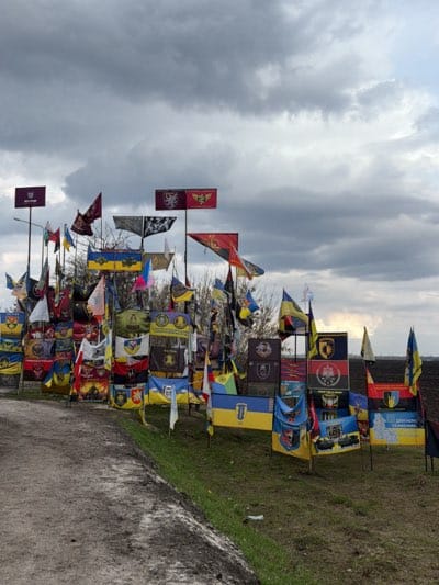 The image shows a memorial or display of flags and banners positioned on a grassy embankment in what appears to be a rural or semi-rural setting. The collection includes Ukrainian national flags as well as flags and banners with various crests, emblems, and insignias.
The background includes an overcast sky, and vegetation, suggesting an outdoor location. The scene's arrangement and visual content suggest a memorial or commemorative site, possibly related to the conflict in Ukraine.
Based on the information available, it is not possible to independently verify the location of "Sad, Ukraine" but given the presence of Ukrainian flags it is likely the photo was taken in the region.